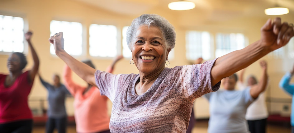 Smiling older woman exercising in a group fitness class, raising her arms with others in the background.
