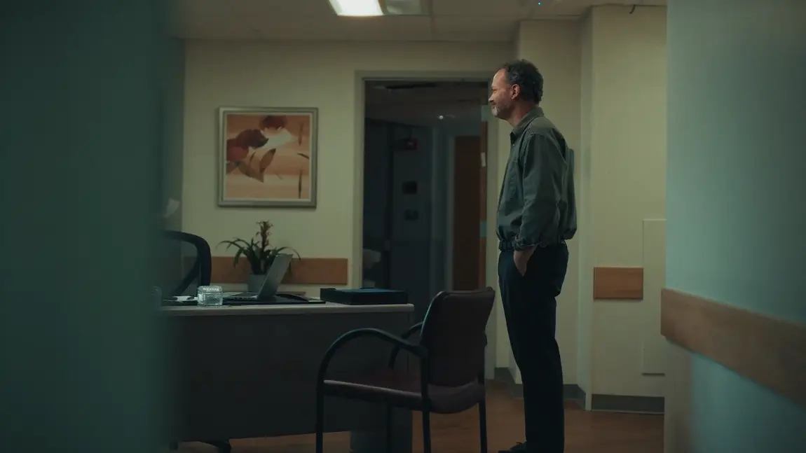 Man stands with hands in pockets in a dimly lit medical office with a desk, chair, and laptop.