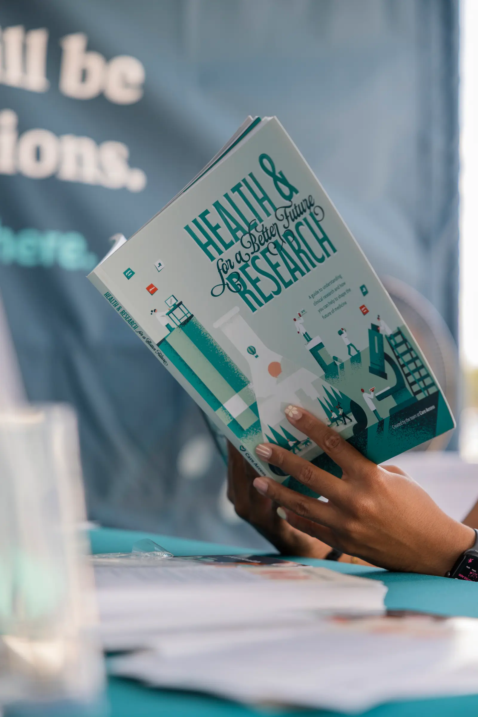 A hand holds open a copy of Health & Research for a Better Future at a Care Access event table.