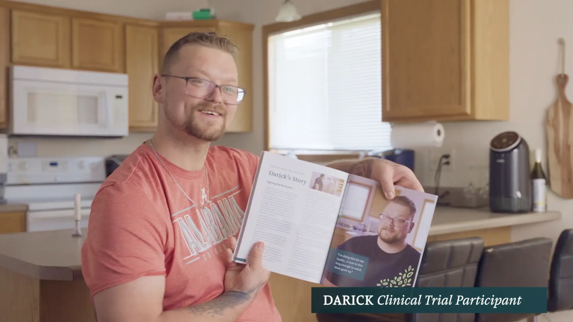 Darick, a clinical trial participant, smiling in his kitchen as he holds open the book to the page telling his story