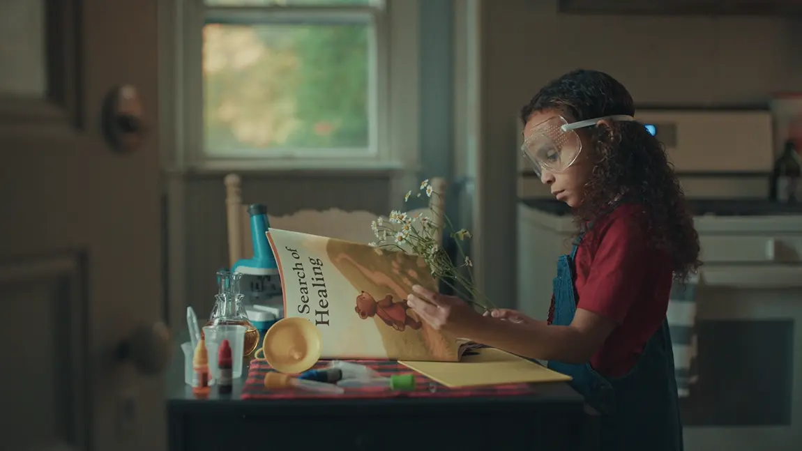 Young girl in safety goggles holds daisies beside an open book and toy science kit on a kitchen table.