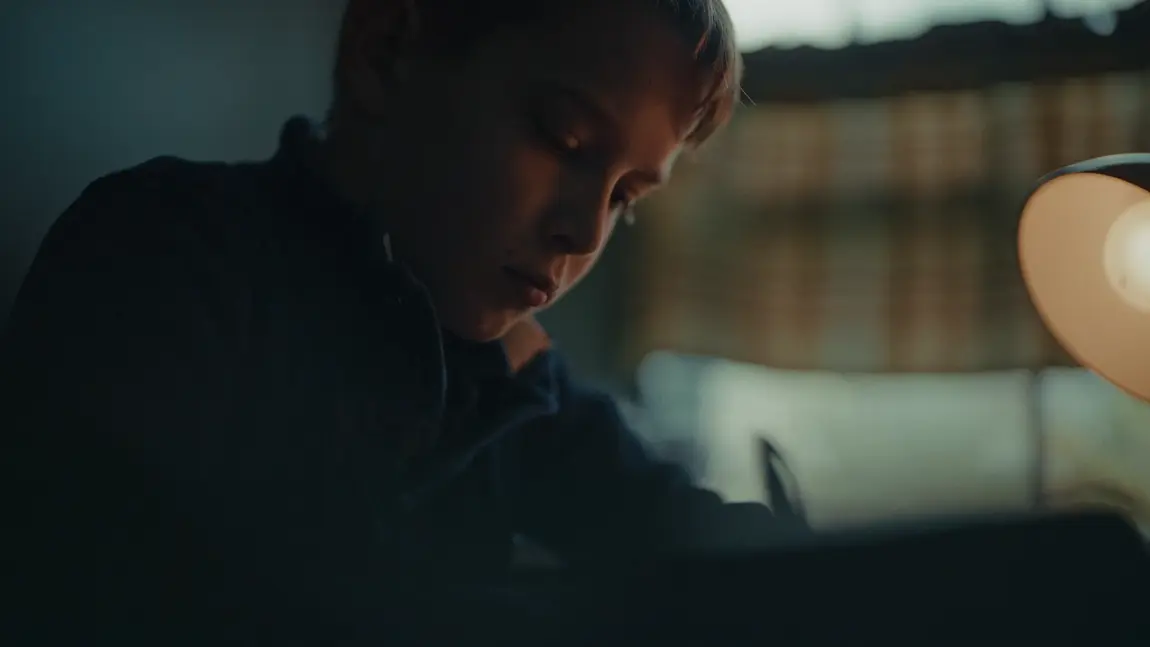 Kid looks down in concentration in a dimly lit room beside a warm desk lamp.